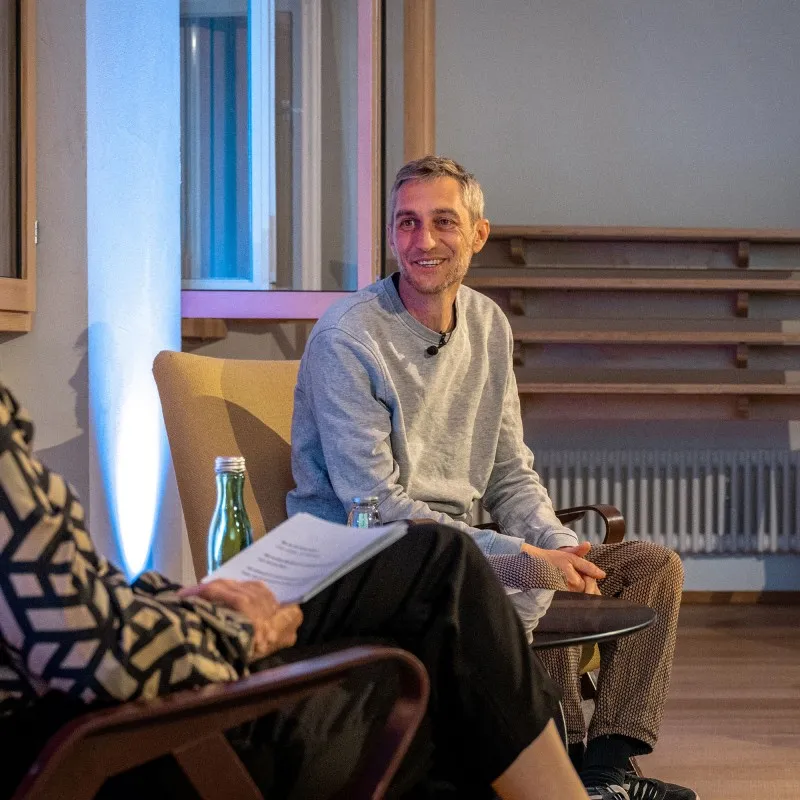 Philipp Böhler (MC Pille One) sitzt bei einer Veranstaltung der ArbeitsLebensGeschichten in der Schaffarei der AK Vorarlberg in Feldkirch auf einem Stuhl und spricht mit einer Moderatorin. Beide sind einander zugewandt, zwischen ihnen steht ein kleiner Tisch mit Wasserflaschen. Die Atmosphäre wirkt ruhig und aufmerksam, der Raum ist modern und warm beleuchtet.