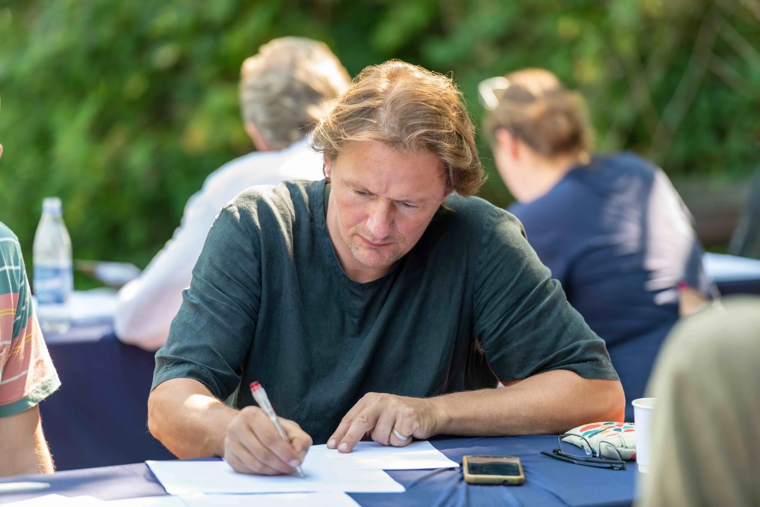 Mann mit hellbraunen Haaren und grünem Pullover schreibt etwas ein Blatt Papier
