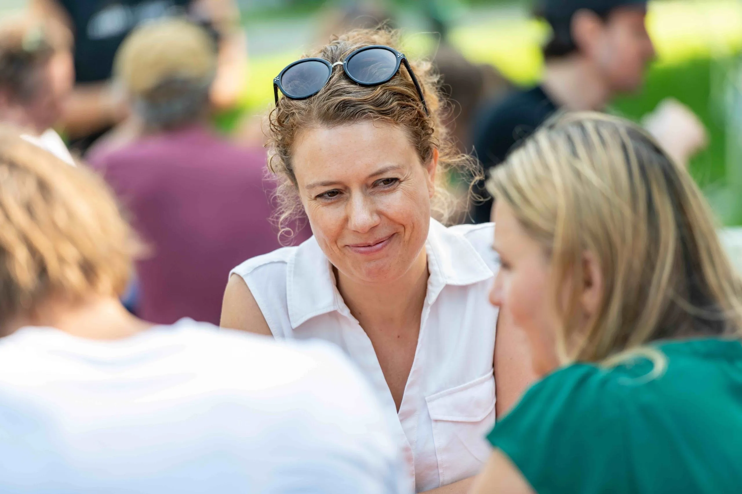 Lächende Frau mit Sonnenbrille im Haar und weißem Oberteil und 2 Frauen sind von hinten zu sehen