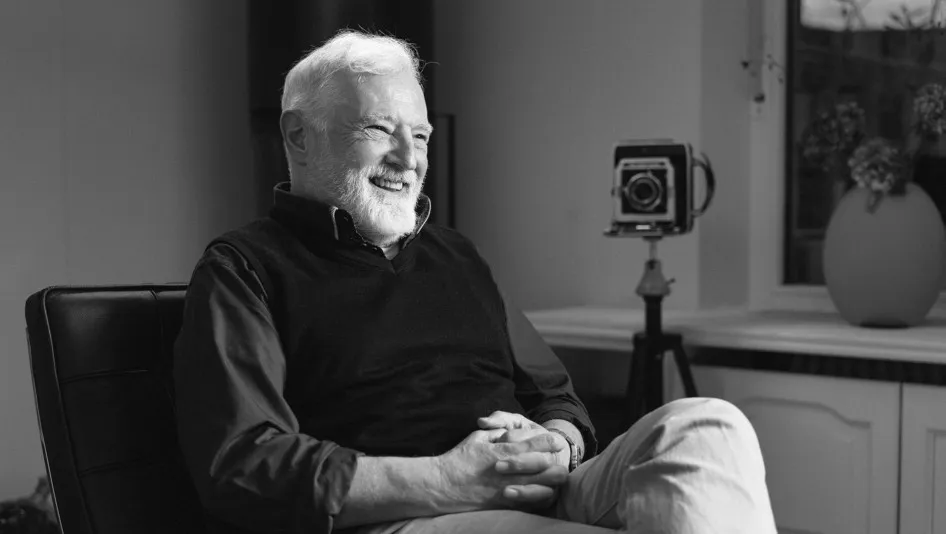 Schwarz-Weiß-Porträt des Fotografen Christian Stuppner: Ein älterer Mann mit weißen Haaren und Bart sitzt entspannt auf einem Stuhl, lächelt und blickt zur Seite. Im Hintergrund steht eine Kamera auf einem Stativ neben einem Fenster und einer Vase.
