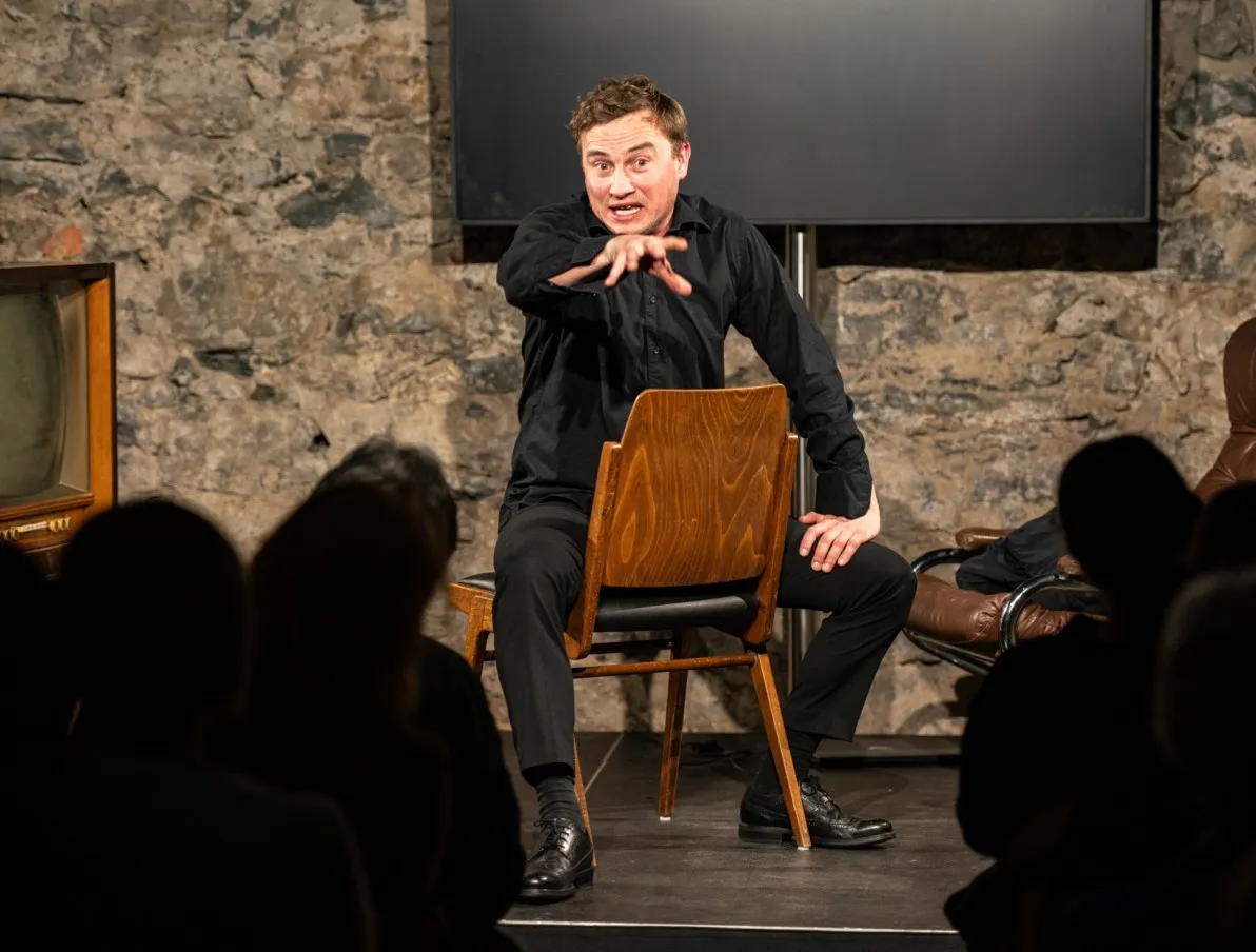 Schauspielszene aus dem Theaterstück „Move fast and break things“ von Daniela Egger, gezeigt im Schaffarei Club in Feldkirch: Ein Schauspieler in schwarzer Kleidung sitzt verkehrt herum auf einem Holzstuhl auf der Bühne und gestikuliert eindringlich mit ausgestreckter Hand ins Publikum. Im Vordergrund sind die Silhouetten der Zuschauer:innen zu sehen, im Hintergrund eine Steinwand, ein Bildschirm und ein Ledersessel.