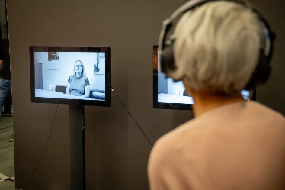 Frau mit Kopfhörer steht seitlich vor einem Bildschirm mit Iris Mähr als Standbild